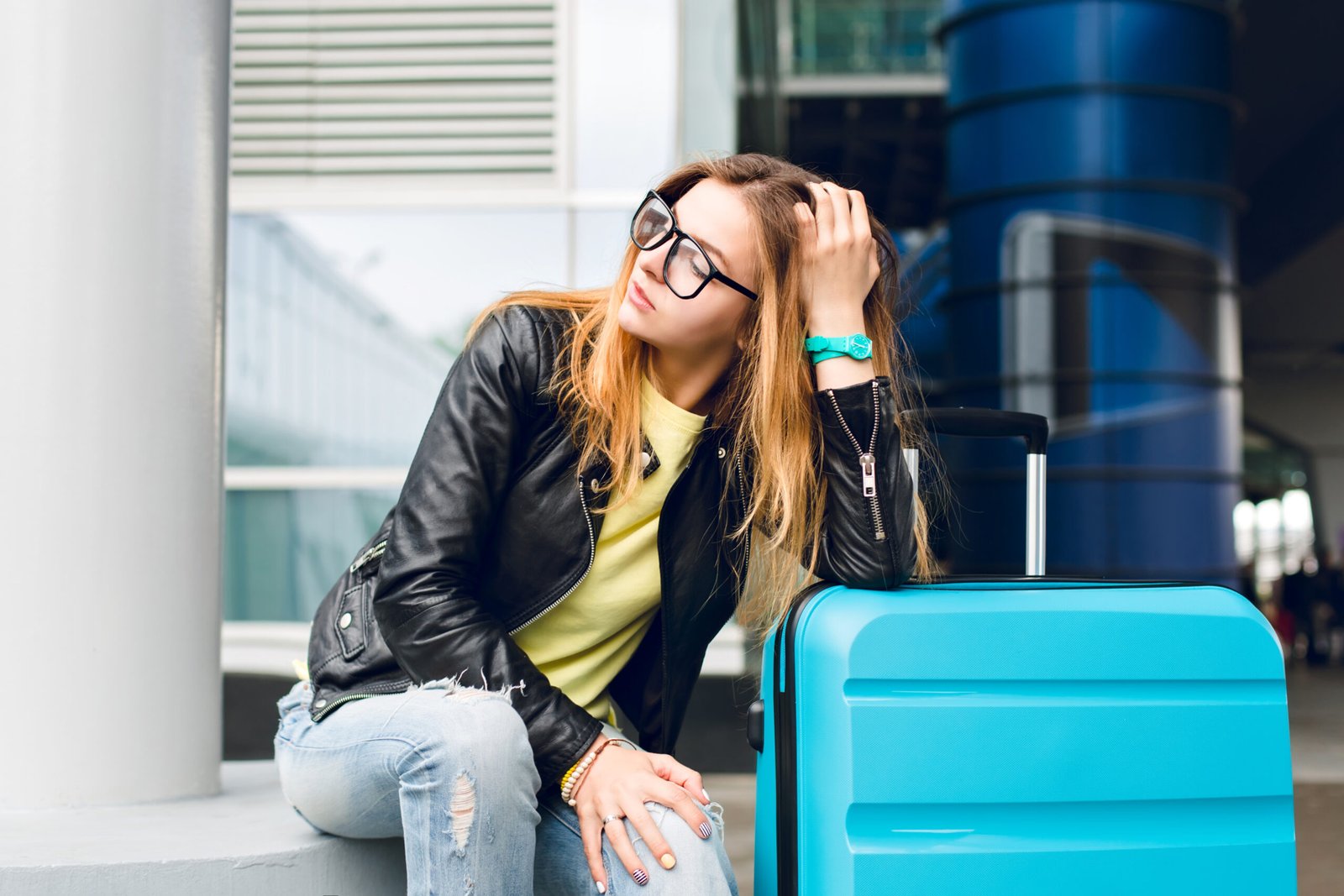 Portrait of pretty girl with long hair in glasses sitting outside in airport. She wears yellow sweater with black jacket and jeans. She leaned to the suitcase and is looking far away
